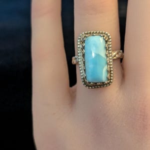 May include: A silver ring featuring a rectangular, light blue, marbled stone set in a beaded bezel. The ring band has a twisted, rope-like texture. The stone displays variations of sky blue and white, creating a smooth, polished surface appearance.