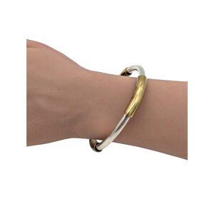 Peut inclure: Un bracelet jonc composé d'une tige argentée lisse agrémentée d'un segment cylindrique doré poli et brillant. Ce bijou présente une esthétique métallique bicolore avec un profil arrondi qui entoure la zone du poignet.