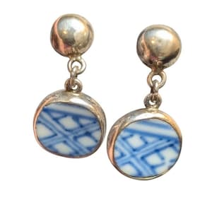 May include: A pair of drop earrings featuring a polished silver-tone ball stud connected to a circular pendant. Each pendant contains a white ceramic insert decorated with a blue geometric cross-hatch pattern set within a smooth silver-tone metal frame.