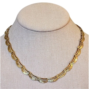 May include: A gold-toned metal necklace featuring a series of repeating, curved, fan-shaped links. Each link has a textured, ridged surface that catches the light, creating a continuous, scalloped pattern that encircles the neck in a uniform design.