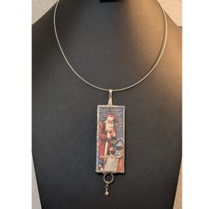 Puede incluir: Un collar rígido de metal plateado con un colgante rectangular que muestra una ilustración de estilo vintage de Santa Claus junto a dos niños en un entorno nevado. El colgante está enmarcado en metal y tiene un pequeño dije circular.