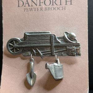 May include: A silver-toned pewter brooch featuring a wheelbarrow, a hat, and garden tools. Two charms hang from the base: a small shovel and a watering can. The packaging is labeled DANFORTH PEWTER BROOCH, DANFORTH PEWTERERS LTD. MADE IN MIDDLEBURY, VERMONT USA.