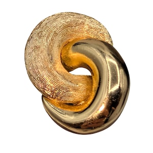 May include: A gold-toned metal brooch featuring two interlocking circular links. One link has a textured, brushed finish with fine concentric lines, while the other link has a smooth, polished, and reflective surface. The design is abstract and circular.