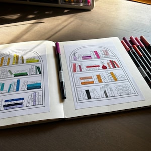 May include: An open notebook with two pages featuring drawings of bookshelves filled with colorful books. A purple pen lies on the left page, and several pens with red and black ink are on the right. The notebook is on a wooden surface.