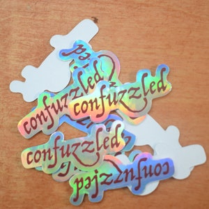 May include: A pile of iridescent holographic stickers with the word "confuzzlled" written in maroon cursive on each sticker.