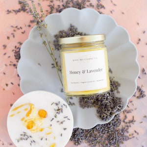 Puede incluir: Una vela de vidrio en un frasco con tapa dorada y una etiqueta blanca que dice "Basic Bee Candle Co." y "Honey & Lavender". La vela está sobre un plato gris con flores de lavanda esparcidas a su alrededor.