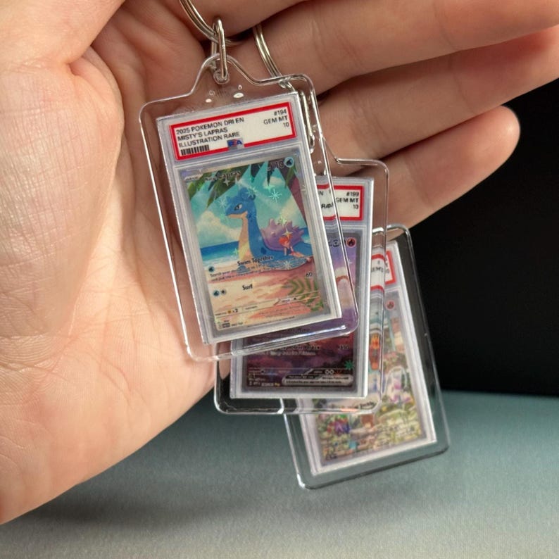 May include: Clear acrylic keychains featuring Pok&eacute;mon trading cards. The keychains display various cards, including a blue Lapras card with text that reads "2025 Pokemon Dr/En Misty's Lapras Illustration Rare". Each card is encased in a protective holder.