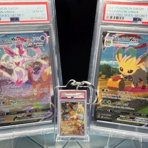 May include: A keychain featuring a graded Pok&eacute;mon card is displayed in front of two other graded Pok&eacute;mon cards. The cards have colorful illustrations of Pok&eacute;mon characters. The keychain card shows a character with orange and white tones.