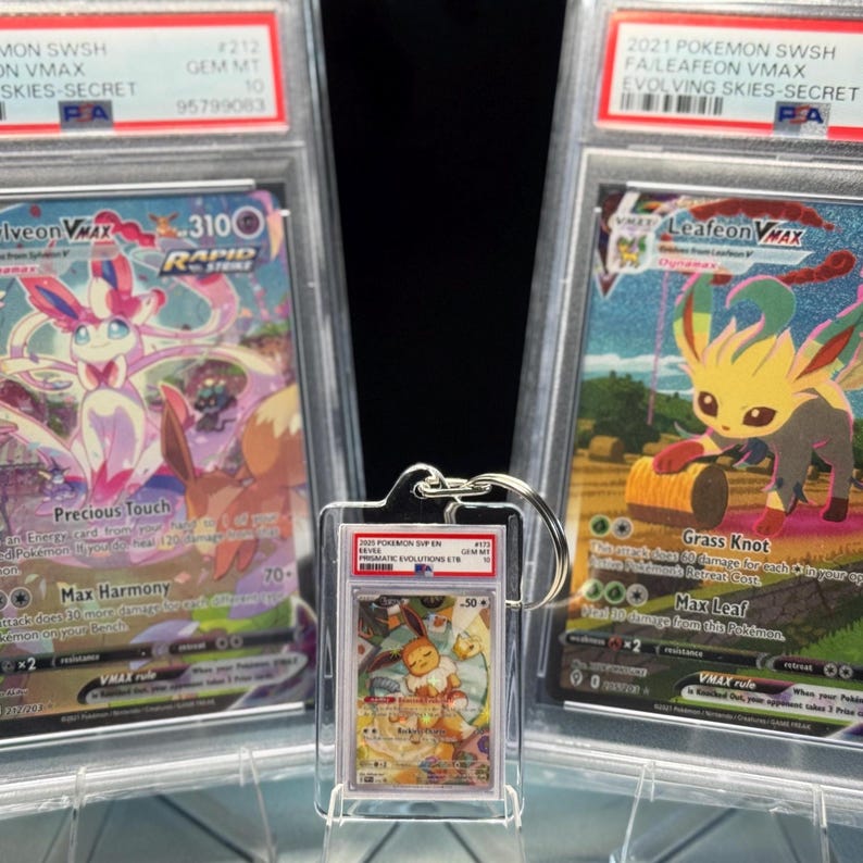 May include: A keychain featuring a graded Pok&eacute;mon card is displayed in front of two other graded Pok&eacute;mon cards. The cards have colorful illustrations of Pok&eacute;mon characters. The keychain card shows a character with orange and white tones.