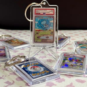 May include: Clear acrylic keychains featuring Pok&eacute;mon trading cards. The central keychain displays a Mew card with a blue and white illustration. Other keychains with various Pok&eacute;mon cards are visible, all encased in protective holders.