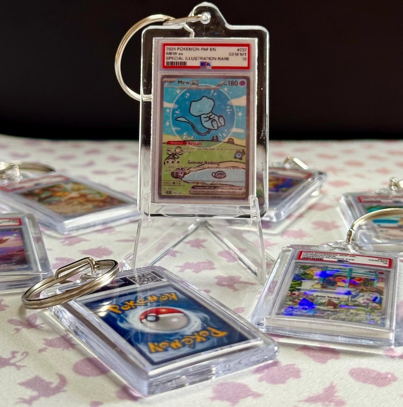 May include: Clear acrylic keychains featuring Pok&eacute;mon trading cards. The central keychain displays a Mew card with a blue and white illustration. Other keychains with various Pok&eacute;mon cards are visible, all encased in protective holders.