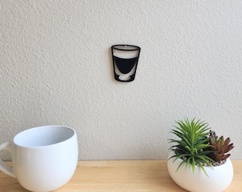 May include: A white ceramic mug and a white ceramic planter with a small succulent plant are on a wooden surface. A black silhouette of a shot glass is on the wall behind the mug and planter.