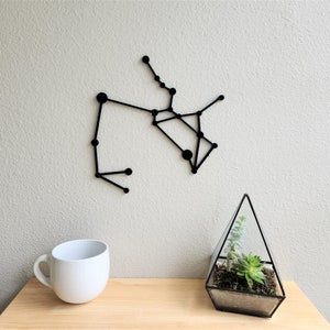 Könnte beinhalten: Schwarze Wanddekoration aus Metall in Form des Sternbildes Schütze. Die Dekoration ist an einer weißen Wand montiert, mit einer weißen Tasse und einem Glas-Terrarium mit einer Sukkulentenpflanze auf einem Holztisch.