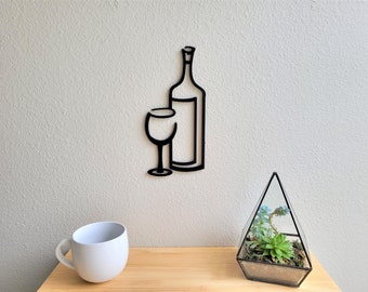 May include: Black metal wall art featuring a wine bottle and glass. The art is mounted on a white wall with a small succulent terrarium and a white mug on a wooden surface below.