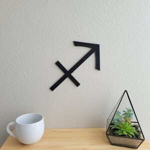 May include: Black wooden Sagittarius zodiac sign on a white wall with a small glass terrarium with succulents on a wooden table.