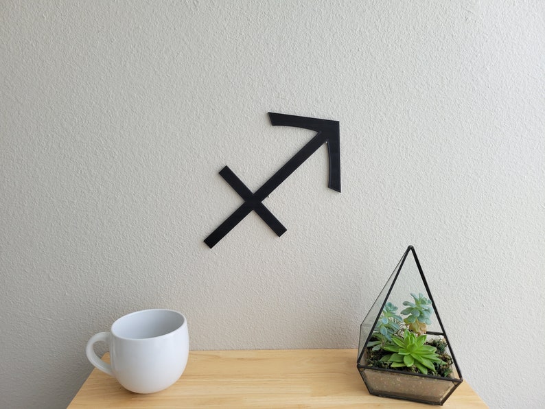 May include: Black wooden Sagittarius zodiac sign on a white wall with a small glass terrarium with succulents on a wooden table.