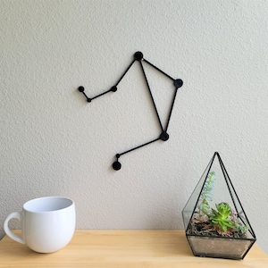 May include: Black metal wall decor in the shape of the Libra constellation. The decor is hanging on a white wall with a white mug and a glass terrarium with a succulent plant on a wooden table.