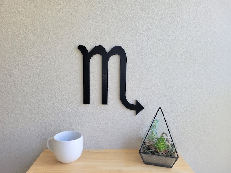 May include: Black metal wall decor in the shape of the Scorpio zodiac sign. The sign is mounted on a white wall. A white mug and a glass terrarium with a succulent plant are on a wooden table in the foreground.