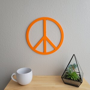 May include: Orange peace sign wall decor with a white mug and a glass terrarium with a succulent plant on a wooden surface.
