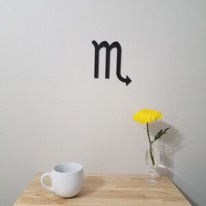 May include: A white ceramic mug sits on a wooden table next to a single yellow flower in a glass vase. A black metal zodiac symbol for Scorpio is on the wall behind the table.