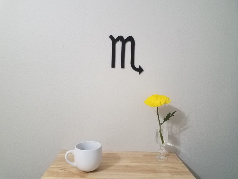 May include: A white ceramic mug sits on a wooden table next to a single yellow flower in a glass vase. A black metal zodiac symbol for Scorpio is on the wall behind the table.