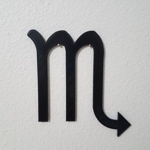 May include: Black metal cutout of the astrological sign Scorpio. The sign is shaped like an 'M' with a tail that curves to the right and ends in an arrow.