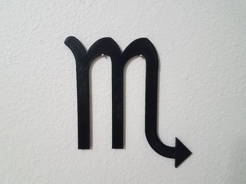 May include: Black metal cutout of the astrological sign Scorpio. The sign is shaped like an 'M' with a tail that curves to the right and ends in an arrow.