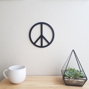 May include: A black peace sign wall decor with a white background. The peace sign is hanging on a white wall. There is a white mug and a glass terrarium with succulents on a wooden table in the foreground.