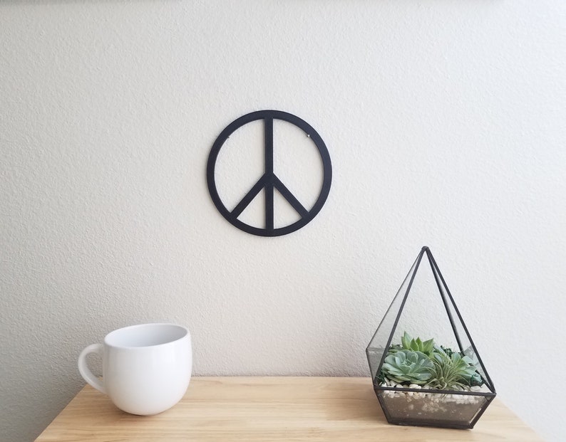 May include: A black peace sign wall decor with a white background. The peace sign is hanging on a white wall. There is a white mug and a glass terrarium with succulents on a wooden table in the foreground.