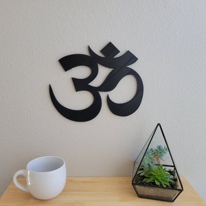May include: Black metal Om symbol wall decor with a white mug and a glass terrarium with green plants on a wooden surface.