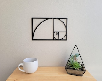 Può includere: Un decoro da parete in metallo nero con la spirale di Fibonacci, nota anche come spirale aurea, una sequenza matematica che appare in natura.