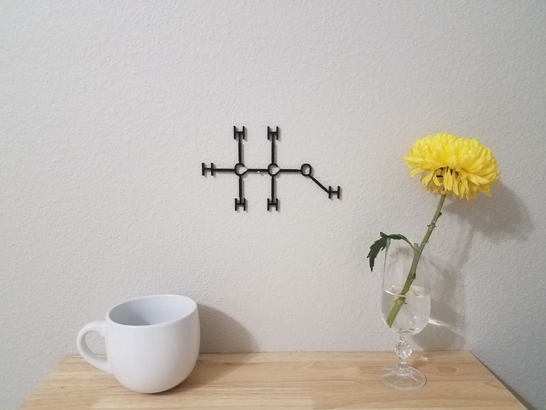 May include: A black metal wall decor in the shape of a chemical structure with the letters 'H' and 'C' on a white wall. A white mug and a yellow flower in a glass vase are on a wooden surface.