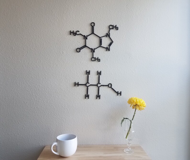 May include: Two black metal wall decor pieces depicting chemical structures. The top piece is a caffeine molecule. The bottom piece is a butane molecule. A yellow flower in a glass vase sits on a wooden surface in front of the wall decor.