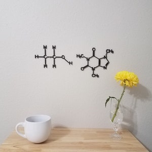 May include: Two black metal wall decor pieces depicting the chemical structures of caffeine and ethanol. The caffeine molecule is on the right and the ethanol molecule is on the left. A white mug and a yellow flower in a glass vase are on a wooden table in the foreground.