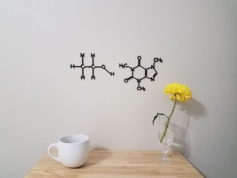 May include: Two black metal wall decor pieces depicting the chemical structures of caffeine and ethanol. The caffeine molecule is on the right and the ethanol molecule is on the left. A white mug and a yellow flower in a glass vase are on a wooden table in the foreground.