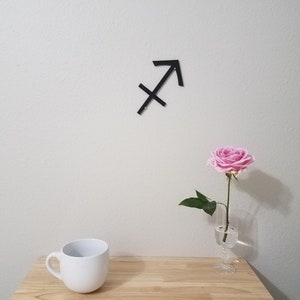 May include: A black metal Sagittarius zodiac sign is mounted on a white wall. A single pink rose is in a clear glass vase on a wooden table. A white ceramic mug is on the table.