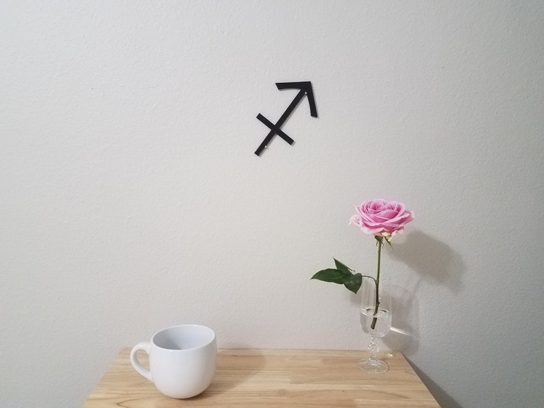 May include: A black metal Sagittarius zodiac sign is mounted on a white wall. A single pink rose is in a clear glass vase on a wooden table. A white ceramic mug is on the table.