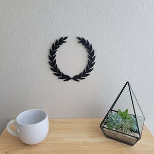 K&ouml;nnte beinhalten: Ein schwarzer Kranz aus Bl&auml;ttern h&auml;ngt an einer wei&szlig;en Wand &uuml;ber einem Holztisch. Eine wei&szlig;e Keramiktasse und ein gl&auml;sernes Terrarium mit Sukkulenten stehen auf dem Tisch.