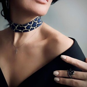 Peut inclure: Un collier ras du cou en perles bleu et blanc avec un motif géométrique. Le collier est porté avec un haut noir et un pendentif en forme de croix argentée. Une bague noire avec un motif de serpent est portée au doigt.