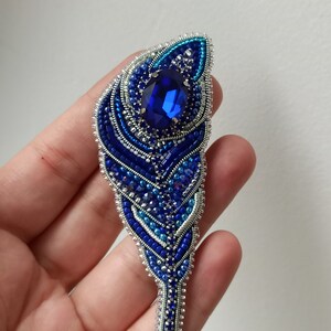 May include: A blue and silver beaded brooch in the shape of a feather with a large blue rhinestone in the center.