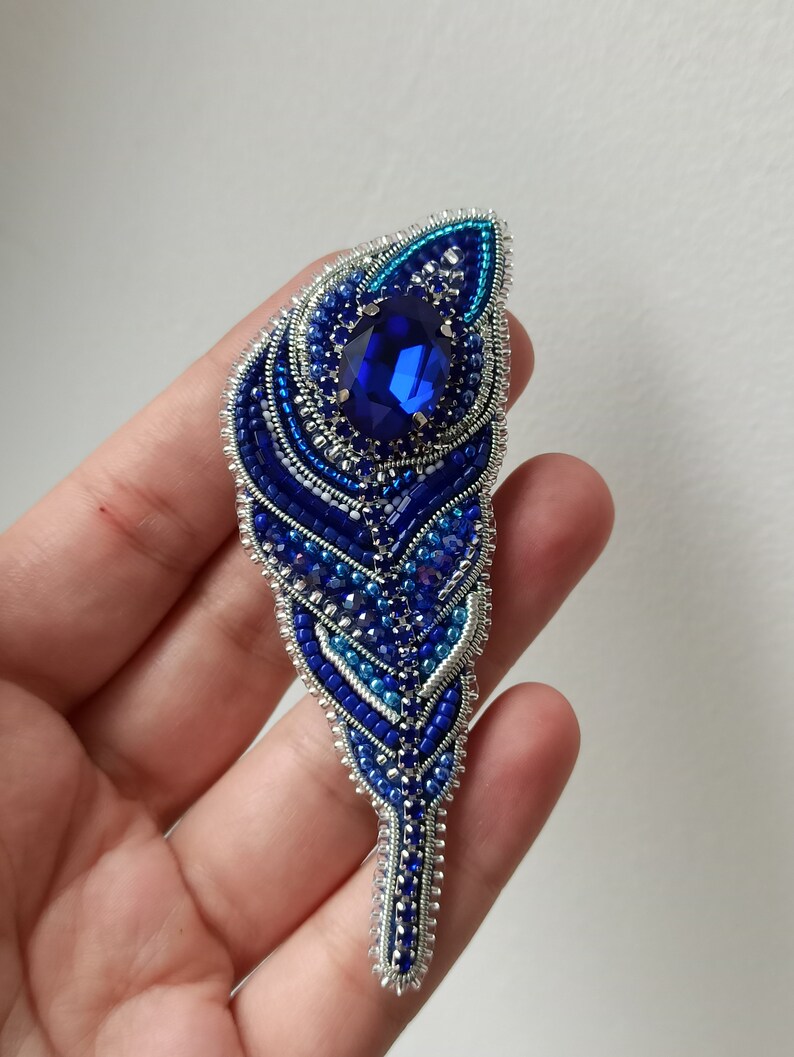 May include: A blue and silver beaded brooch in the shape of a feather with a large blue rhinestone in the center.