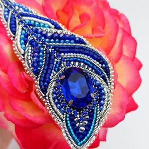 May include: A blue and silver beaded brooch with a large blue gemstone in the center. The brooch is on a pink rose.