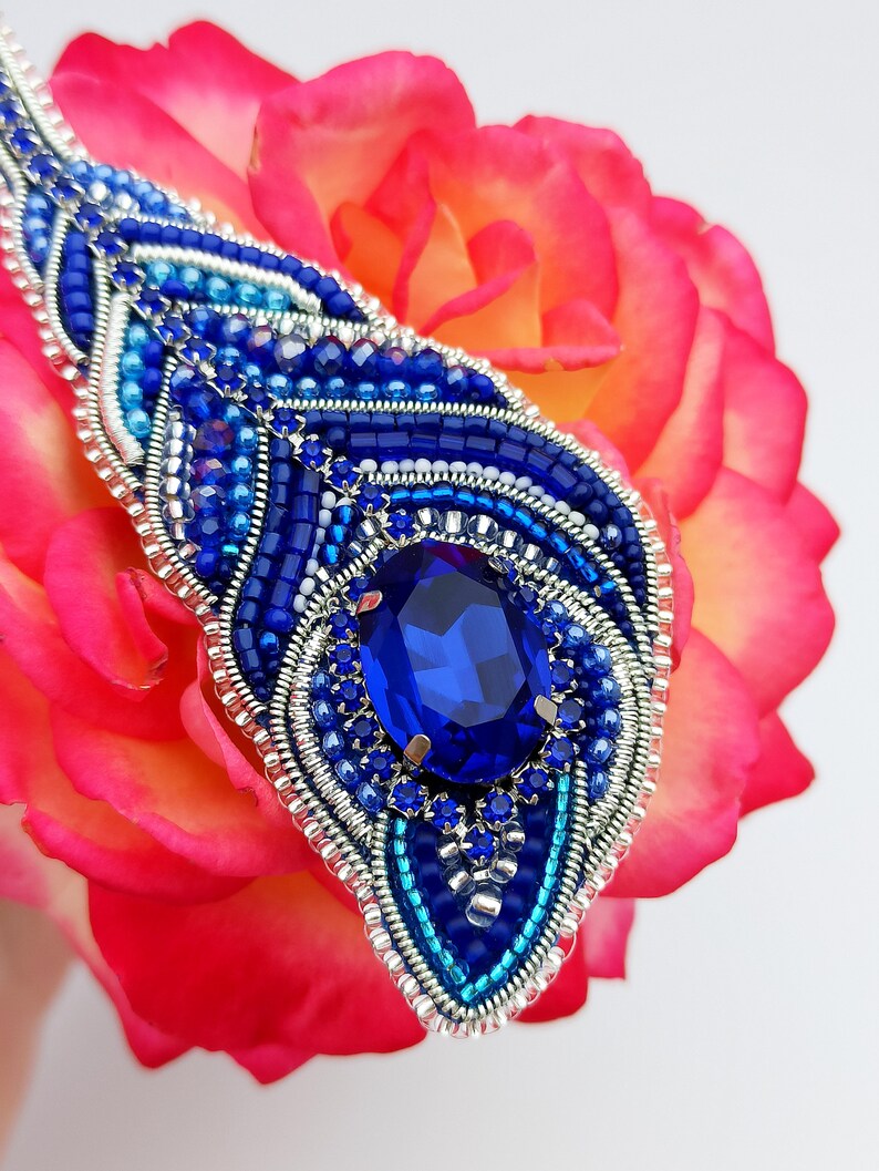 May include: A blue and silver beaded brooch with a large blue gemstone in the center. The brooch is on a pink rose.