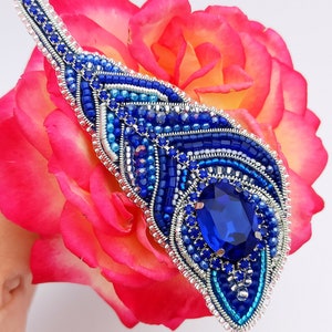 May include: A blue and silver beaded brooch with a large blue gemstone in the center. The brooch is shaped like a feather and is resting on a pink rose.