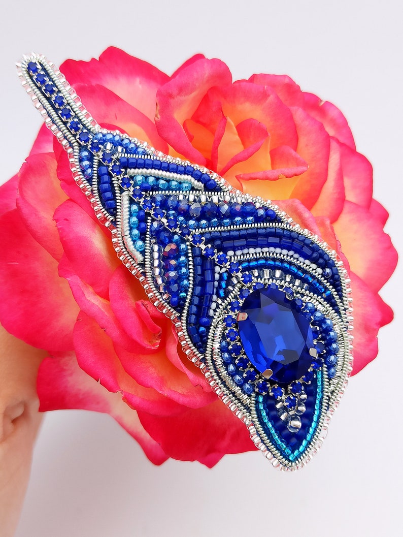 May include: A blue and silver beaded brooch with a large blue gemstone in the center. The brooch is shaped like a feather and is resting on a pink rose.