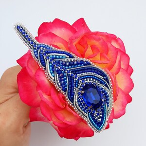 May include: A blue and silver beaded brooch with a large blue gemstone in the center. The brooch is shaped like a teardrop and is set against a pink rose.