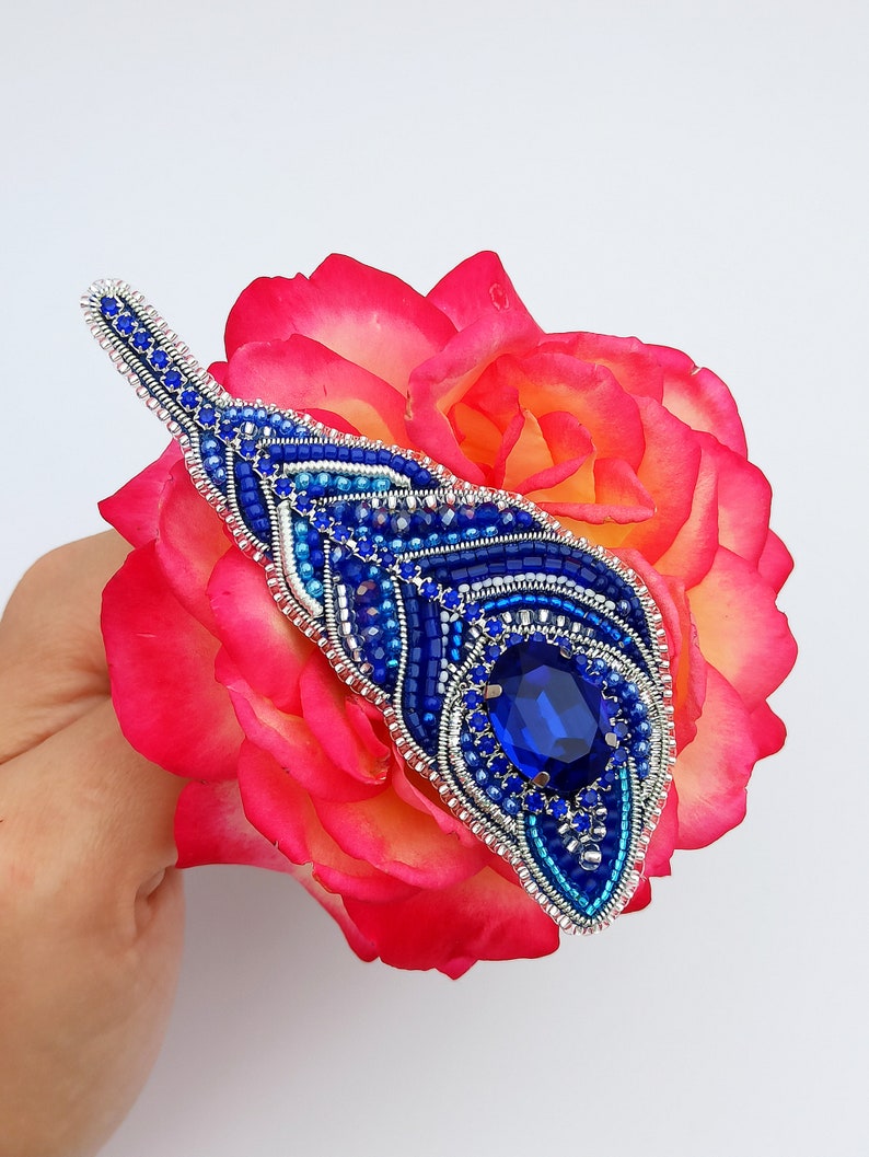 May include: A blue and silver beaded brooch with a large blue gemstone in the center. The brooch is shaped like a teardrop and is set against a pink rose.