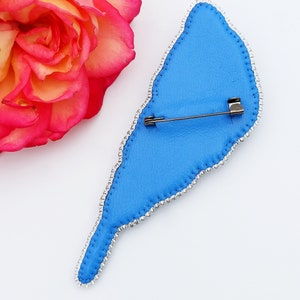 May include: Blue beaded bird brooch with a silver pin on the back. The brooch is shaped like a bird with a long tail.
