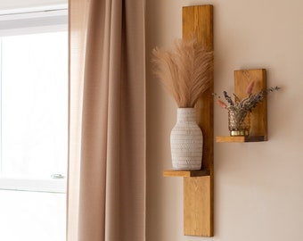 May include: A wooden wall shelf with two tiers, featuring a white ceramic vase with dried pampas grass and a small glass vase with dried flowers. The shelf is mounted on a beige wall with a brown curtain in the background.