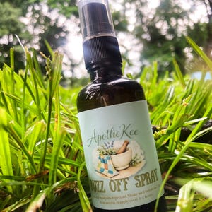 Puede incluir: Una botella de vidrio marrón de "Buzz Off Spray" con un pulverizador negro, colocada en hierba verde. La etiqueta dice "ApotheKee" e incluye una ilustración de mortero y maja. El producto es un repelente de mosquitos.